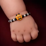 •A child's beaded bracelet worn on the wrist, featuring alternating black and silver beads with an orange "cartoon face" focal point.
